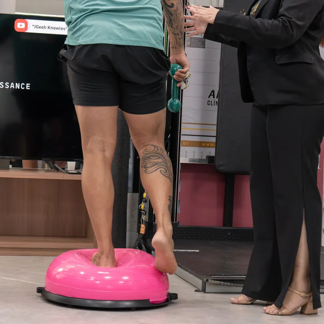 Paciente se equilibra descalço sobre bosu rosa enquanto fisioterapeuta orienta o exercício de propriocepção
