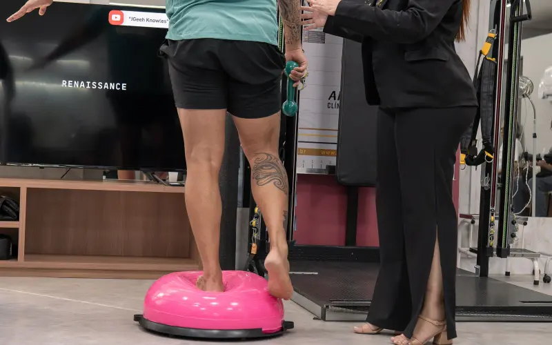 Paciente se equilibra descalço sobre bosu rosa enquanto fisioterapeuta orienta o exercício de propriocepção