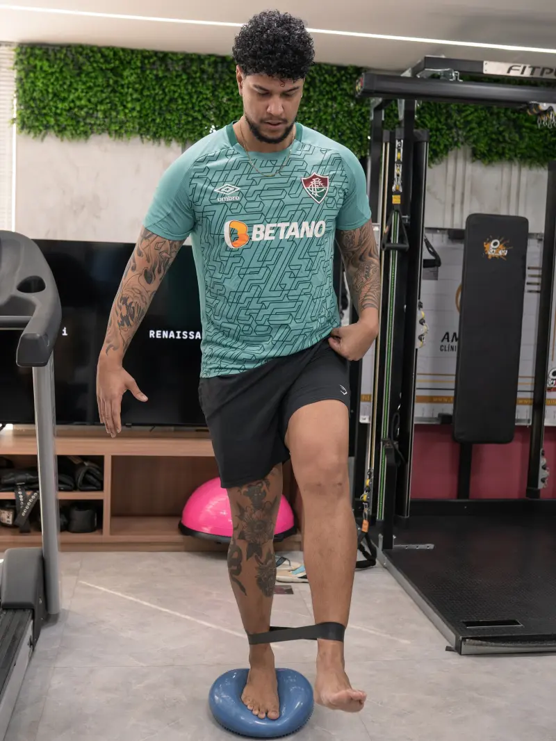 Atleta de futebol se equilibra em um pé sobre disco azul com faixa elástica nos tornozelos
