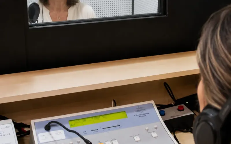Profissional opera o audiômetro enquanto paciente usa fones dentro da cabine acústica
